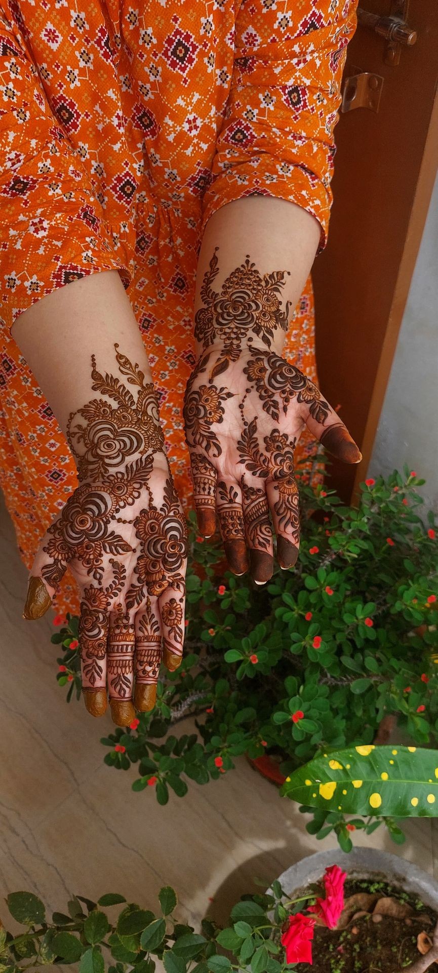 Hands with intricate mehndi designs, wearing an orange patterned dress, surrounded by potted plants.