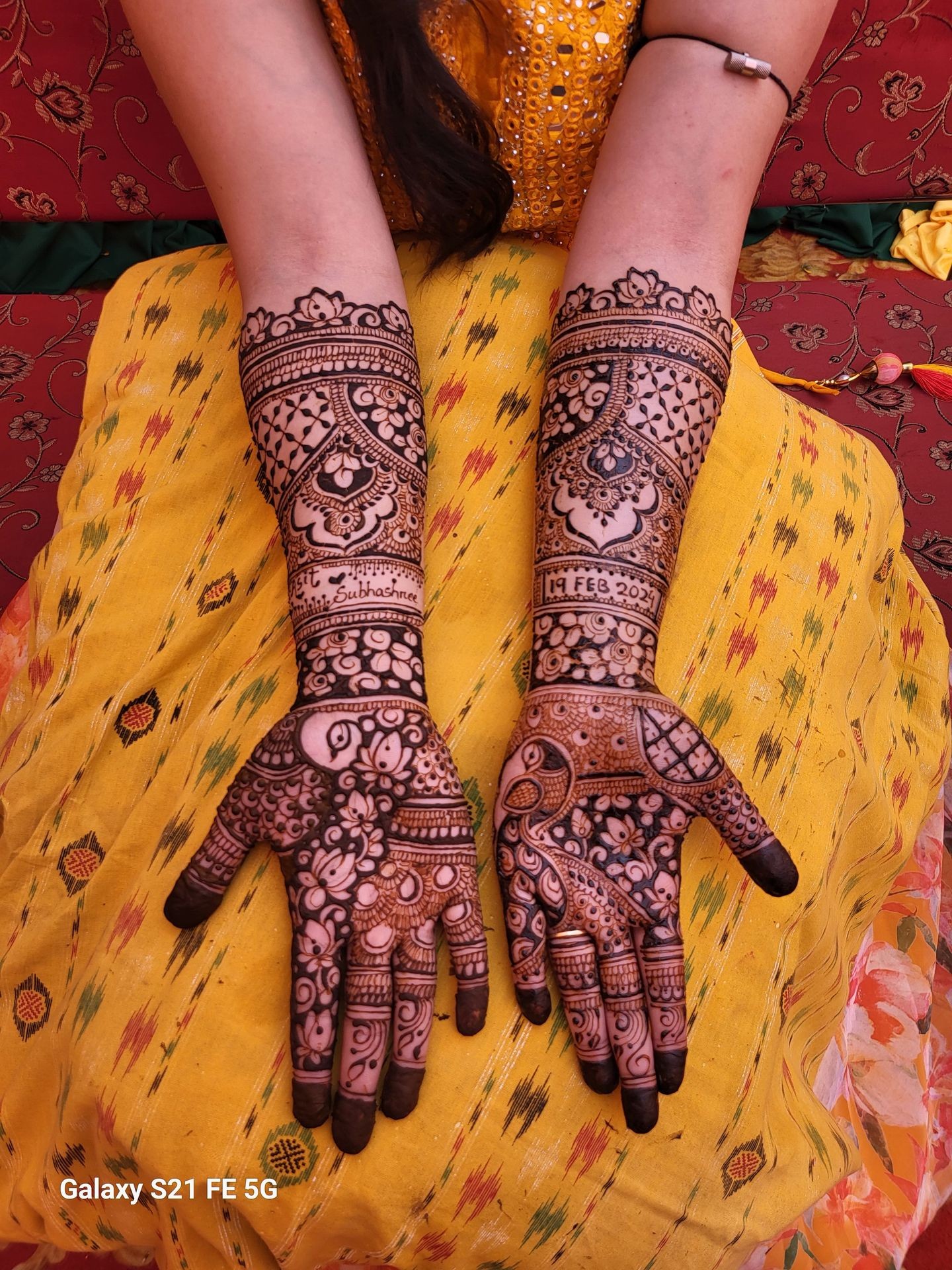 Hands with intricate henna patterns on forearms, wearing a yellow dress.